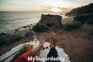 En la imagen se ven los pies de una pareja al borde de la costa en una colina. Observan el atardecer y disfrutan de un trago. Compartir tiempo es muy importante para una relación. Por eso, regalar esto seria el regalo adecuado. 