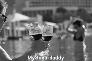 Sugar Dating: Vacaciones con el Sugar Daddy Sugar Dating: En la imagen vemos a un hombre sosteniendo dos copas de vino. La imagen esta en blanco y negro. En el primer plano se encuentra el hombre que se puede ver de forma clara y precisa. Es mayor de edad, ya que tiene pelo blanco. En el fondo, se puede ver una mujer saliendo de una piscina. Esta desenfocada, pero parece querer entrar en contacto con este hombre.
